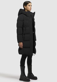 Melnas pūkainās mētelis ar kapuci, ar kvadrātveida šuvēm, priekšējo rāvējslēdzēju, sānu kabatām un vidēju garumu, uzvilkts virs melnajiem legingiem un apaviem.