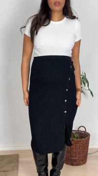 Žena odjevena u bijelu majicu kratkih rukava, crnu midi suknju s gumbima i prorezom te crne čizme do koljena stoji unutra pokraj tkane košare za biljke.