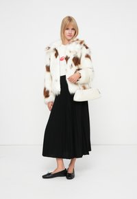 Veste en fausse fourrure blanche avec des taches brunes, un tee-shirt graphique, une jupe plissée noire et des ballerines noires. Sac à main blanc avec une forme arrondie et une poignée.