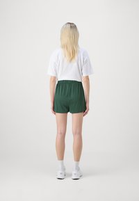 Zöld sportnadrág rugalmas derékkal, sima fehér pólóval párosítva. A jelenet minimalista, semleges háttérrel.