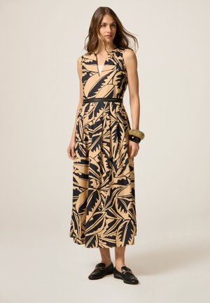 Maxi dress in beige with black leaf patterns, sleeveless design, V-neckline, cinched waist, and a flowing skirt. Accented with a black belt.
