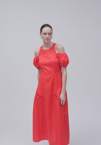Femme debout dans une robe rouge à épaules dénudées avec des manches bouffantes et des détails froncés près des hanches sur un fond gris uni.