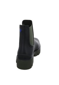 Botín negro de goma con secciones laterales elásticas verdes y una suela texturizada. Presenta un acento azul cerca del talón.