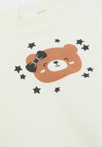 Langärmliger cremefarbener Sweatshirt mit einem braunen Bären-Motiv und einer schwarzen Schleife, umgeben von schwarzen Sternen. Aus weichem Baumwollmaterial, schlichtes Design.