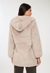 Cappotto beige con cappuccio realizzato in materiale fluffy, presenta una vestibilità comoda, maniche lunghe e una texture morbida senza chiusure visibili o dettagli.