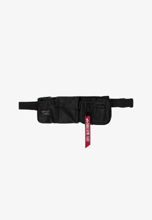 Sac banane noir avec plusieurs poches zippées et à rabat, sangle réglable, et une étiquette en tissu rouge indiquant "REMOVE BEFORE FLIGHT."