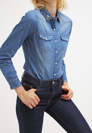 Femme portant une chemise en jean ajustée bleu clair avec un col orné et un jean taille haute bleu foncé, mains reposant près des poches.