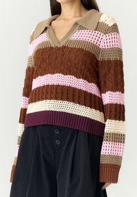 Femme portant un pull en maille multicolore avec des rayures horizontales marron, rose, beige et violettes et un col en V, associé à un pantalon noir.