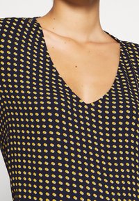 Marinblus med djup V-hals och gula ovala prickar. Slät textur, figurnära design som framhäver halslinningen och mönstret.