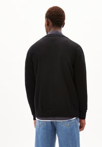 Zwarte gebreide trui met een ronde halslijn, lange mouwen en geribde manchetten, gedragen over een gelaagd blauw gestreept shirt en lichtblauwe jeans.