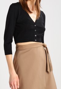 Svart kort cardigan med V-ringning och fem knappar, tillsammans med en beige omslagsskjol med knytning i midjan.