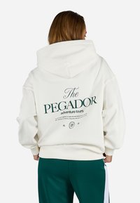 Felpa con cappuccio oversize di colore crema con un grande logo verde e testo sulla schiena. La parte anteriore presenta un design semplice. Tessuto morbido e strutturato, vestibilità casual.