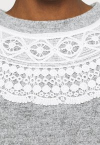 Šedý svetr s bílou krajkovou yoke s složitými vzory a vlnitými okraji. Měkká textura s kulatým výstřihem.
