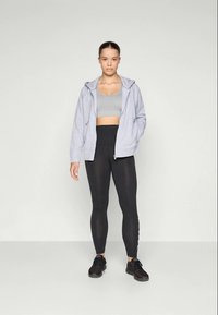 Jovem mulher em pé, vestindo um casaco com fecho cinza, soutien desportivo cinza, leggings pretas e ténis pretos contra um fundo simples.