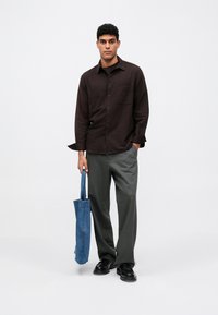 Camicia abbottonata marrone scuro con taschino sul petto, abbinata a pantaloni grigi a gamba larga. Tiene una borsa a tracolla in denim blu. Scarpe nere.