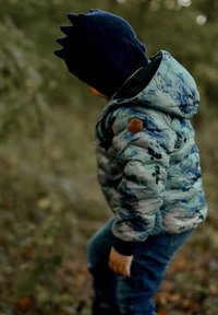 Polsterēta jaka zilā un zaļā abstraktā rakstā, ar kapuci, teksturētu audumu un brūnu sešstūra formālu logo uz pleca.