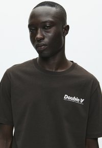 Pruun puuvillane t-särk, mille vasakus rinnapiirkonnas on valge "Double V" logo, klassikalise ümmarguse kaeluse ja lühikeste varrukatega.