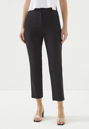 Zwarte, klassiek gesneden broeken met een hoge taille, verticale plooien en een cropped lengte. De stof lijkt glad en zacht, gecombineerd met transparante sandalen met een hak.