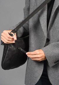 Bolso de cuero negro con correa para el hombro y detalle de cremallera, sobre un blazer gris. Presenta una textura suave y un diseño elegante.