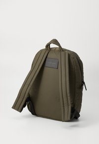 Sac à dos matelassé olive avec un design élégant, comprenant des bretelles rembourrées, une poignée supérieure et un patch logo à l'arrière. Fermeture éclair.