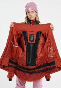 Rote wasserdichte Winterjacke mit gestepptem Design, Netzfutter und mehreren Innentaschen. Eine rosafarbene Mütze wird auf dem Kopf des Models getragen.