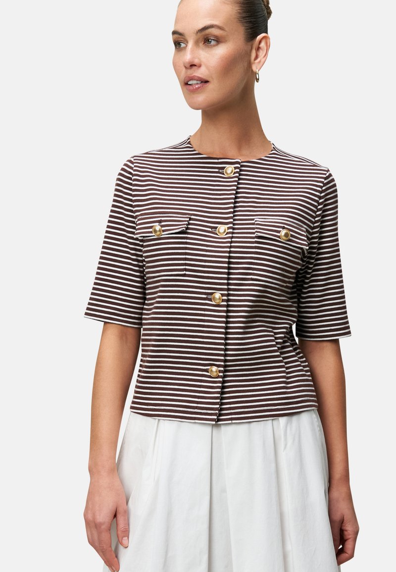 Blusa a maniche corte a righe marroni e bianche con bottoni dorati e due tasche sul petto. Abbinata a una gonna bianca.