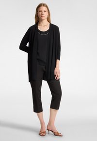 Cardigan lungo nero sopra una maglia nera, abbinato a pantaloni cropped neri e sandali con tacco marroni. L'outfit presenta un design semplice.