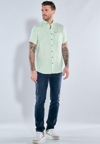 Hellgrünes Kurzarmhemd mit Knopfleiste und Brusttasche, kombiniert mit dunkelblauen Jeans und beigen Sneakers. Glatte Stoffqualität.