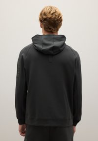 Felpa grigia scura realizzata in tessuto morbido, con cappuccio regolabile, polsini a costine e un design elegante con una toppa sulla spalla sinistra.