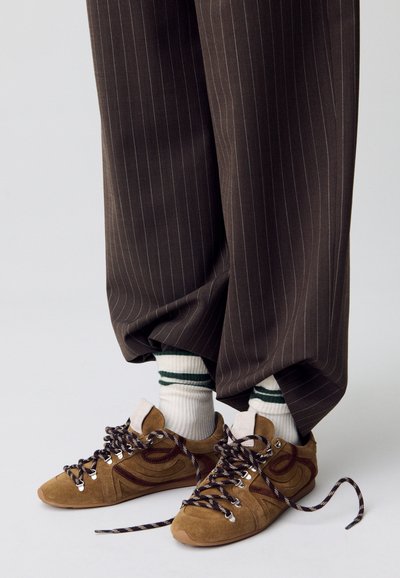Sneaker in suede marrone con dettagli bianchi e lacci a fantasia, abbinati a calzini a righe verdi e bianchi, e pantaloni larghi a righe scure.