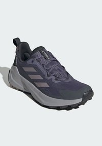 Chaussure de sport violette avec une tige en maille, un motif texturé, trois rayures grises, une coque de protection renforcée et une semelle extérieure épaisse en caoutchouc noir.