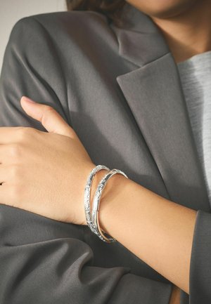 Nahaufnahme einer Person, die zwei silberne, gravierte Armbänder am Handgelenk trägt, gekleidet in einem grauen Blazer und einem hellgrauen Oberteil.