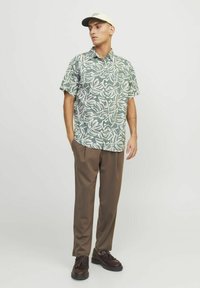 Maglietta a maniche corte con motivo a foglie verdi e bianche, abbinata a pantaloni marroni e scarpe nere, con un cappellino di colore chiaro.