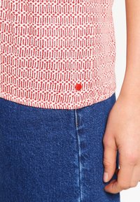 Rød og hvid mønstret top med rund kant, parret med blå denimjeans. Har et lille broderet logo på skjortens nederste kant.