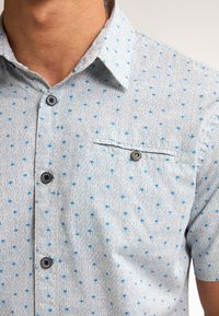 Camicia azzurro chiaro a maniche corte con un sottile motivo testurizzato. Presenta piccoli motivi blu, una tasca sul petto e bottoni scuri.