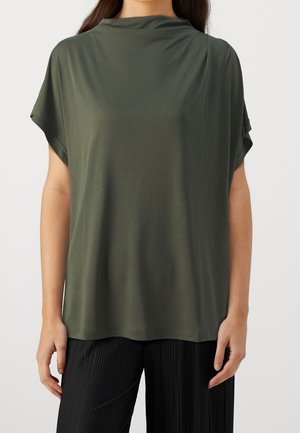 Frau trägt ein locker sitzendes, olivgrünes kurzärmeliges Oberteil mit hohem Ausschnitt und schwarze, plissierte Hose, steht vor einem einfarbigen Hintergrund.