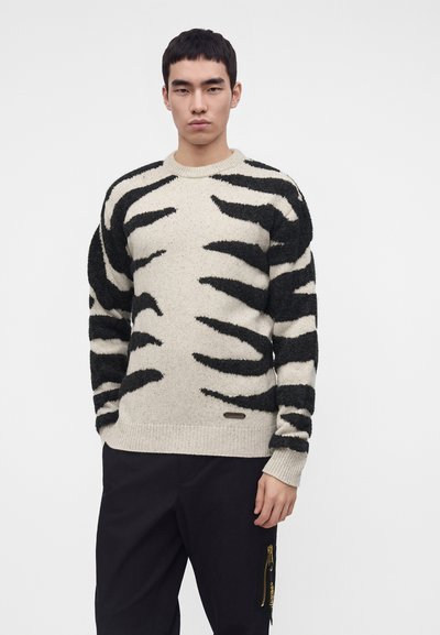 Jeune homme portant un pull beige avec des rayures abstraites de tigre noires et un pantalon noir, debout devant un fond blanc uni.