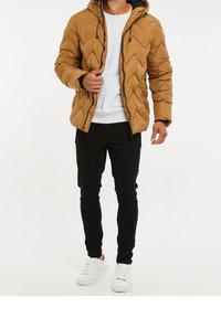 Bruine donsjas met een chevron gestikt patroon, verstelbare capuchon, zwarte rits, gedragen over een grijs T-shirt, gecombineerd met zwarte broek en witte sneakers.