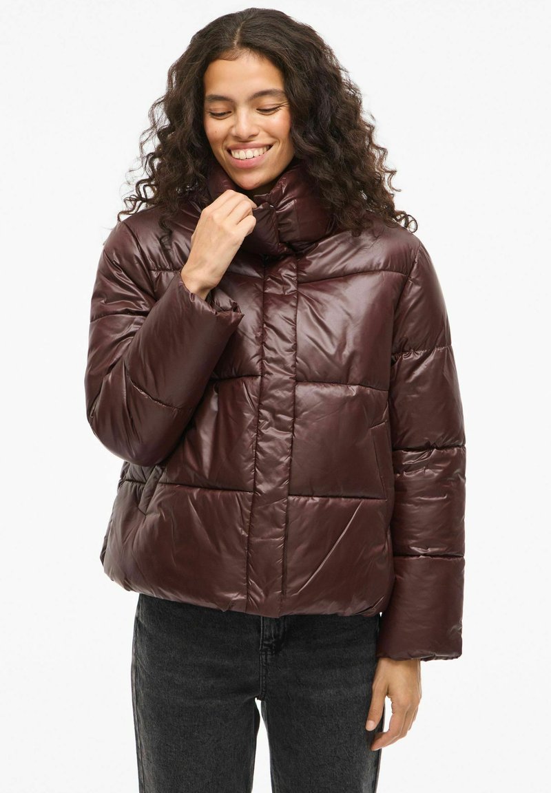 Kratka smeđa puffer jakna s visokim ovratnikom. Izrađena je od sjajnog, quilted materijala. Ima kvadratni kroj i velike šivane panele.