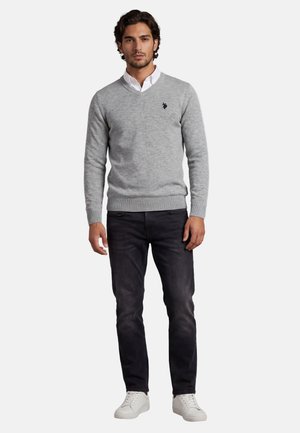 Mann trägt einen hellgrauen Pullover mit V-Ausschnitt über einem weißen Hemd mit Kragen, schwarze Jeans und weiße Turnschuhe, steht vor einem einfachen Hintergrund.