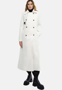 Manteau long blanc avec un design à double boutonnage, de grands boutons et un col large. Porté avec des bottines noires et un pantalon fuselé.