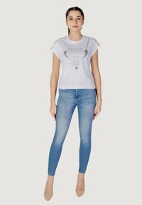 T-shirt bianco in cotone con grafica metallica argento con il testo "GUESS FOLLOW YOUR DESTINY", abbinato a jeans skinny blu chiaro e tacchi beige.