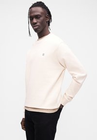 Φόρμα ανοιχτό μπεζ με στρογγυλή λαιμόκοψη, με μικρό μπλε λογότυπο "II" στο στήθος. Μαλακή υφή με μακριά μανίκια και άνετη εφαρμογή.