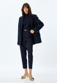 Blazer navy de gran tamaño con botones dorados, suéter ajustado navy, pantalones entallados a juego y tacones dorados tipo slingback con acentos negros.