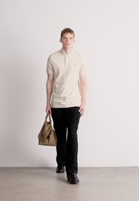 Μπεζ πόλο μπλούζα με διακριτικό λογότυπο, μαύρο παντελόνι και καφέ τσάντα χειρός. Το μοντέλο περπατάει σε ανοιχτό γκρι δάπεδο μπροστά σε λευκό απλό φόντο.