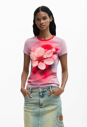 Femme portant un t-shirt rose avec un grand motif de fleur rose et des gouttelettes d'eau, associé à une jupe en denim clair avec des poches.