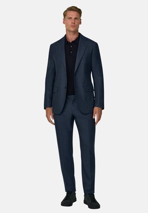 Homme portant un costume bleu foncé avec un polo noir et des baskets noires, debout devant un fond clair uni.