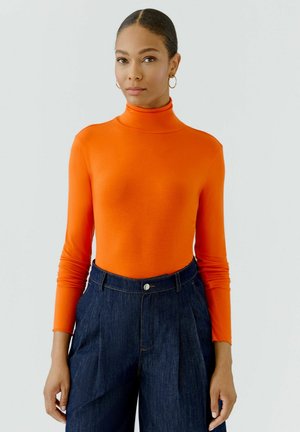 Femme aux cheveux foncés attachés, portant des boucles d'oreilles créoles dorées, un col roulé orange à manches longues et un jean foncé taille haute, debout devant un fond uni.