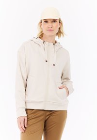 Beige rits-hoodie met een gecentreerde naad, trekkoordcapuchon en kangoeroezakken. Gecombineerd met tan broek en een lichte pet.