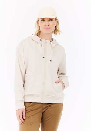 Beige rits-hoodie met een gecentreerde naad, trekkoordcapuchon en kangoeroezakken. Gecombineerd met tan broek en een lichte pet.